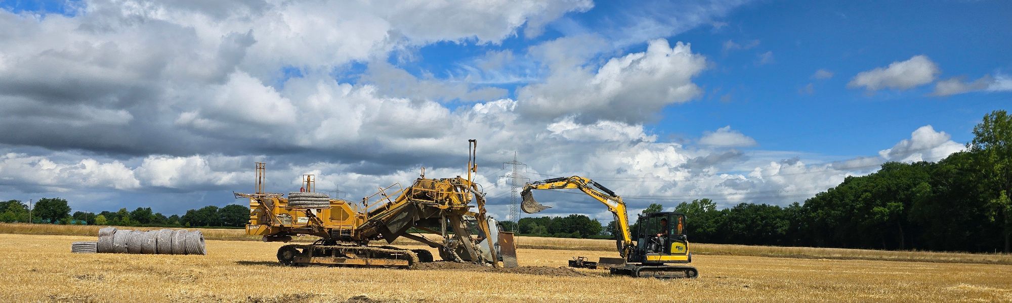 Zu sehen sind unsere Drainagemaschine sowie unser Bagger im Einsatz auf einer landwirtschaftlichen Fläche.